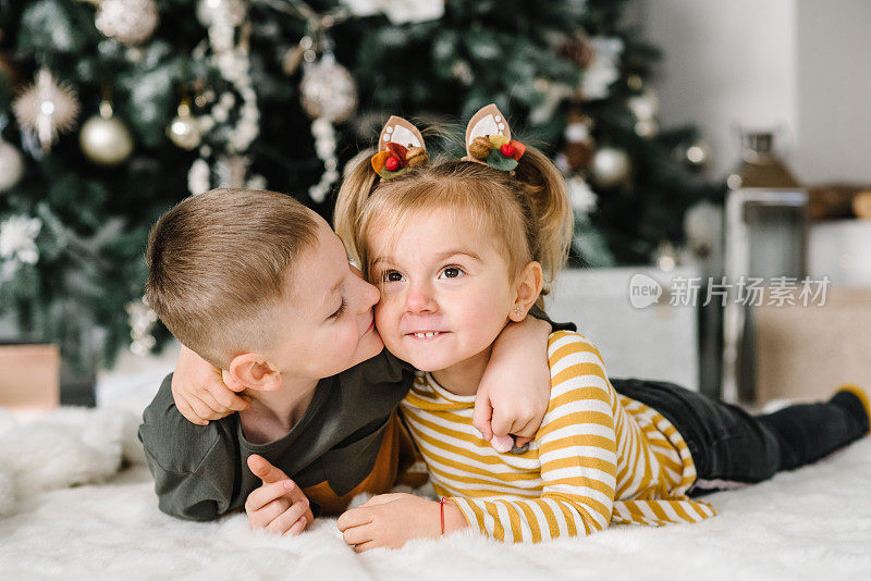 Happy children hugging at a home. Brother kiss sister. Childs playing near Christmas tree. Happy New Year and Merry Christmas. Ð¡oncept of a family holiday. Decorated interior room of a house. Closeup.